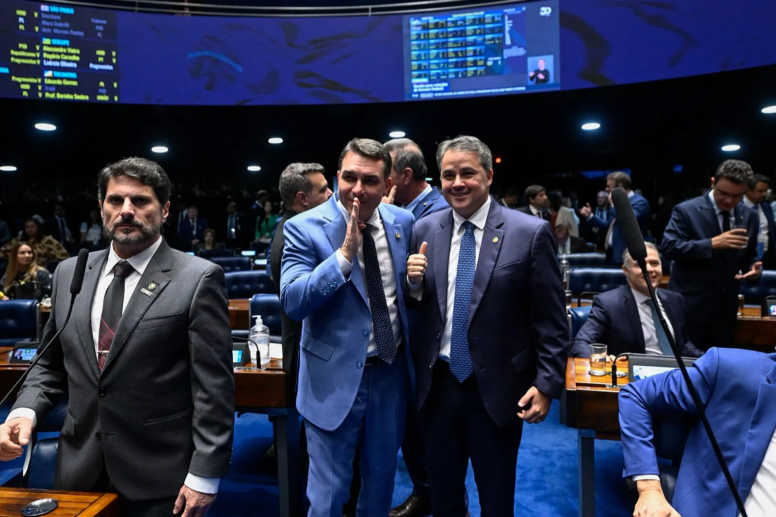 No Plenário do Senado, Flávio Bolsonaro, de terno azul-claro, conversa com o senador Efraim Filho, que faz sinal de positivo; à esquerda, o senador Marcos do Val.