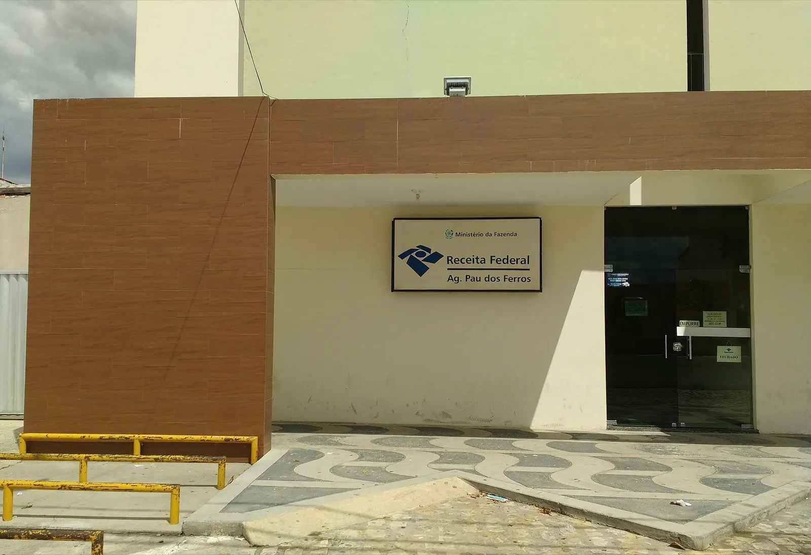 Fachada de uma agência da Receita Federal, com a placa do órgão sobre a entrada, em uma rua de cidade do interior.