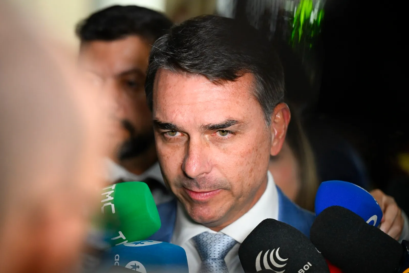 Close de Flávio Bolsonaro, de terno e gravata, diante de vários microfones de emissoras durante entrevista.