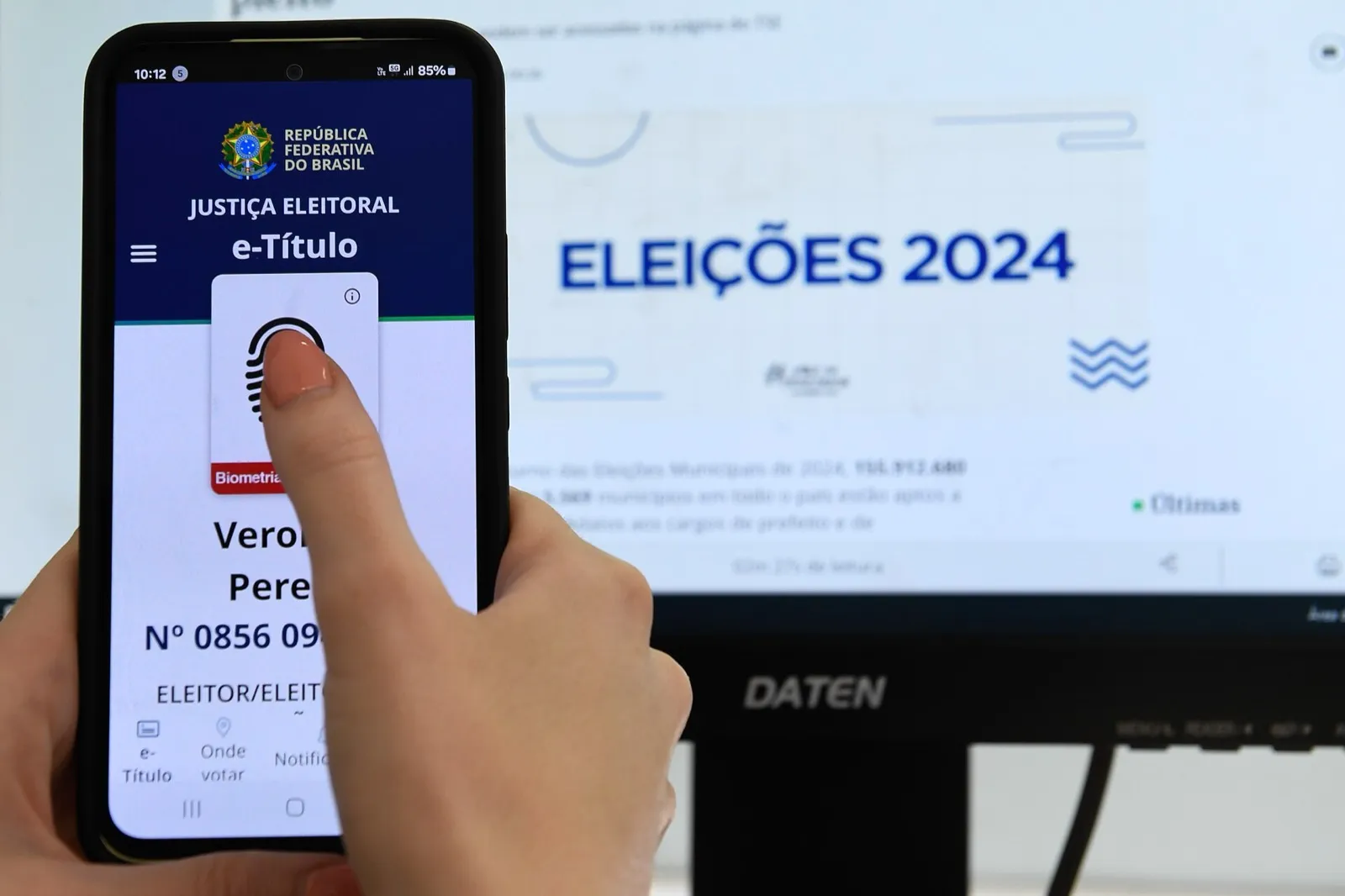 Mão segura um celular com o aplicativo e-Título da Justiça Eleitoral aberto, diante de um monitor com a inscrição Eleições 2024.