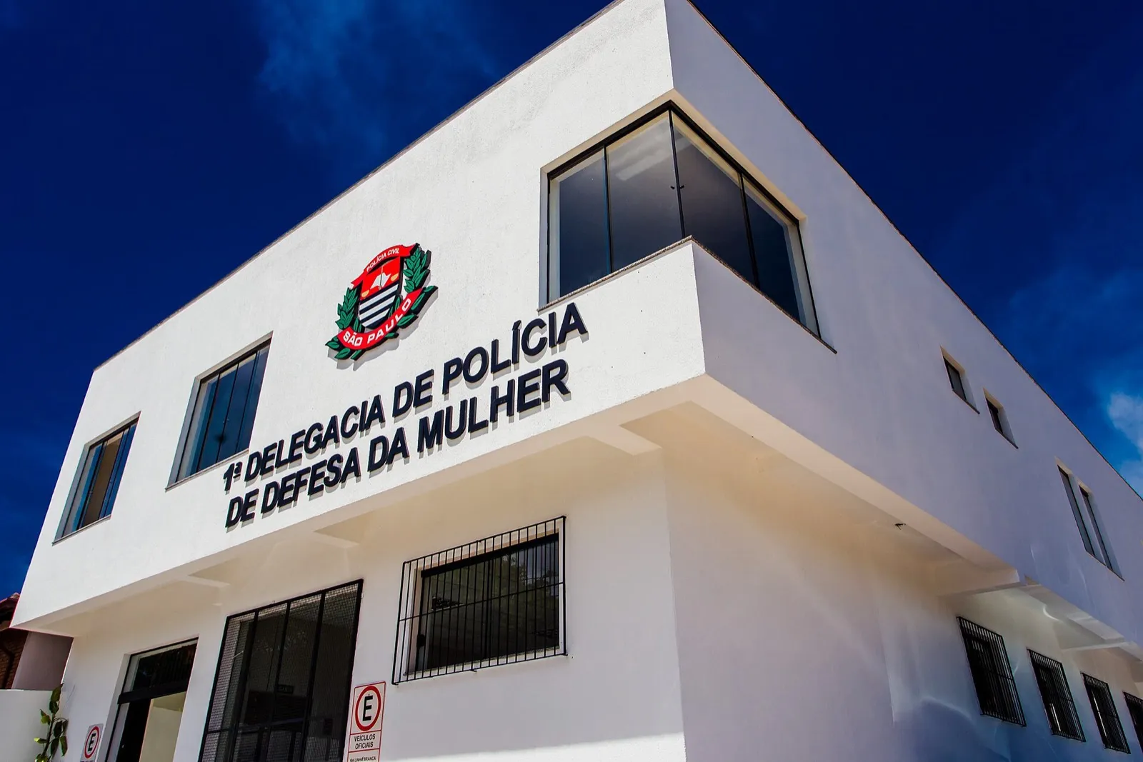 Fachada branca da 1ª Delegacia de Polícia de Defesa da Mulher, com o brasão da Polícia Civil de São Paulo, sob céu azul.