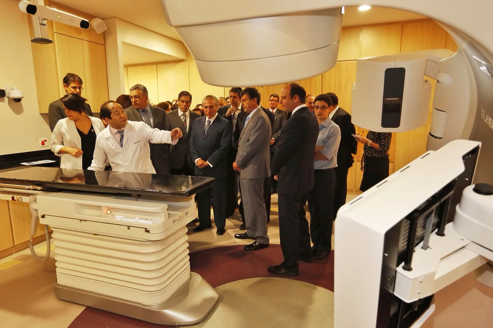 Grupo de homens de terno e profissionais de jaleco observa um equipamento de radioterapia em uma sala de hospital.