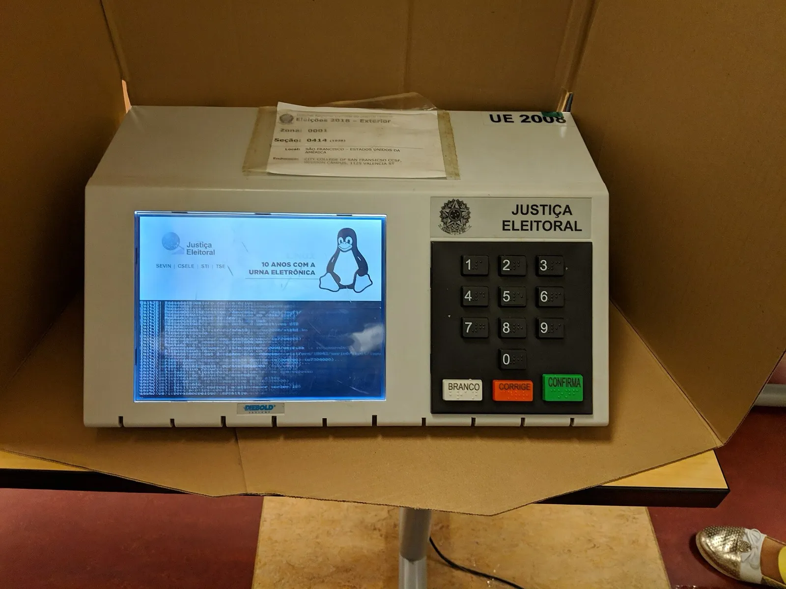 Urna eletrônica modelo UE2008 dentro da cabine de votação, com a tela de inicialização acesa e o teclado numérico com as teclas Branco, Corrige e Confirma.