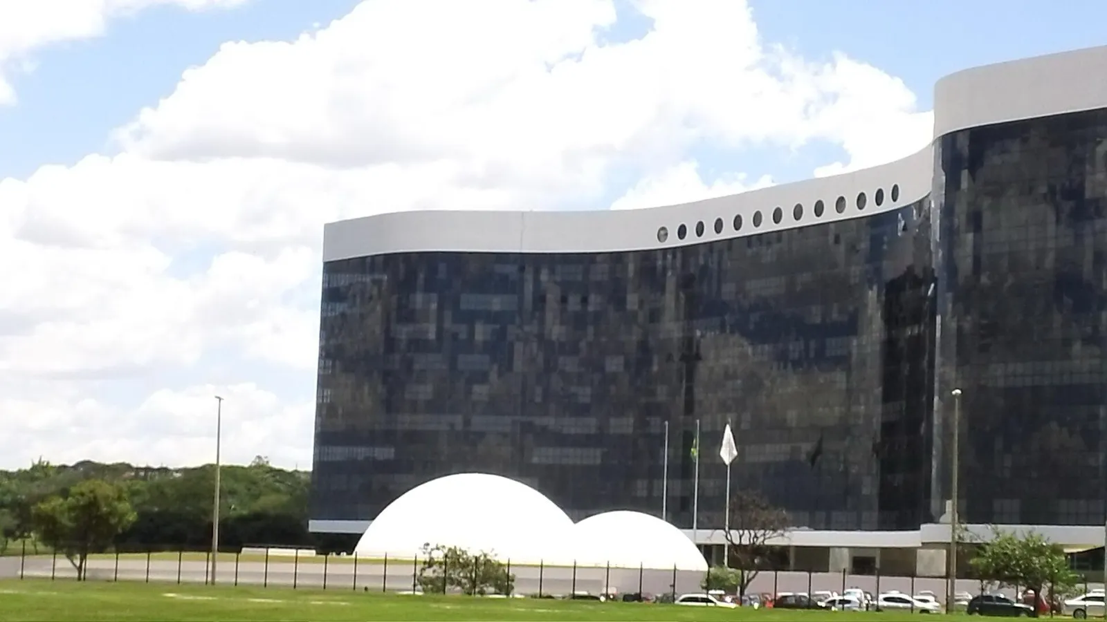 Fachada curva de vidro escuro da sede do Tribunal Superior Eleitoral, com duas cúpulas brancas à frente e céu com nuvens.