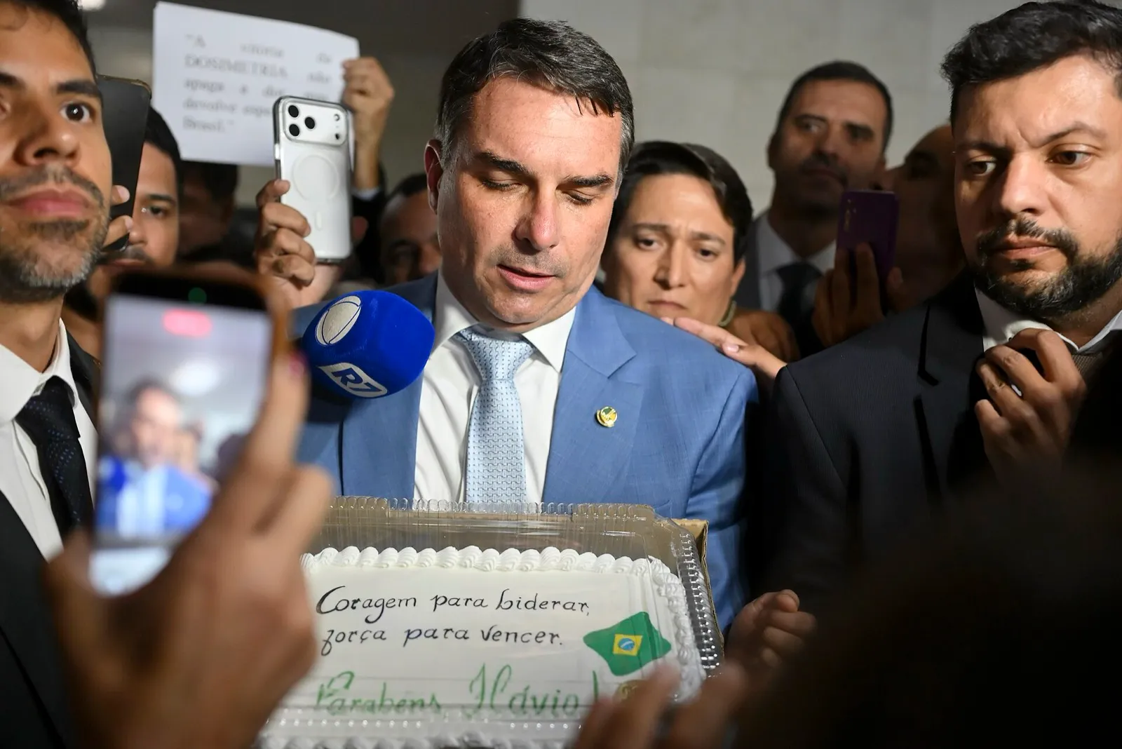 Flávio Bolsonaro, de terno azul, cercado por apoiadores, celulares e um microfone, segura um bolo com a inscrição 'Parabéns Flávio'; ao fundo, um cartaz cita a dosimetria.