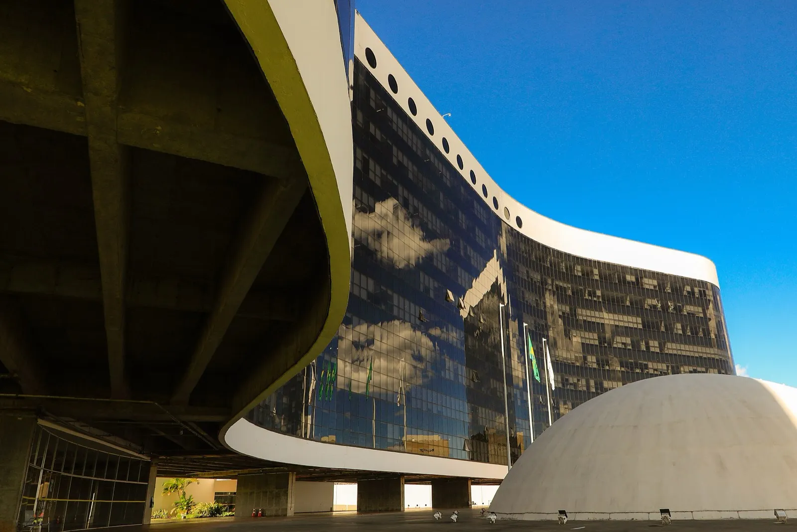 Prédio da sede do Tribunal Superior Eleitoral, com fachada curva de vidro espelhado e cúpula branca, visto sob uma marquise de concreto.