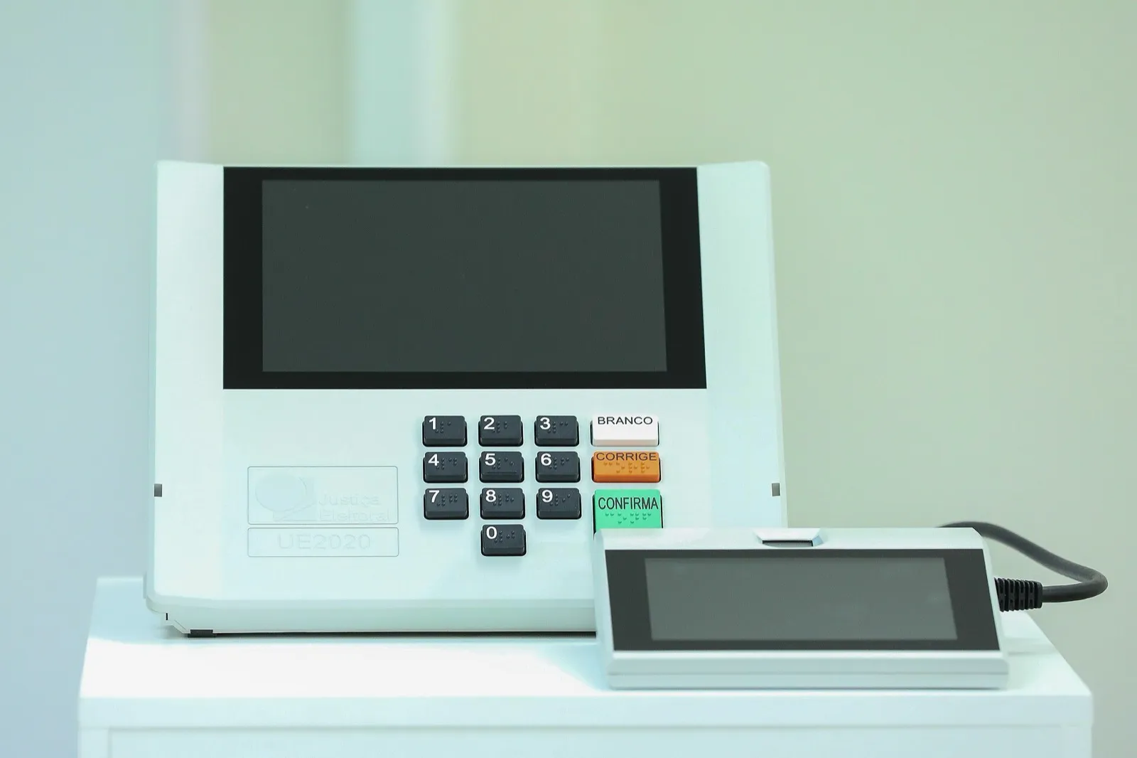 Urna eletrônica branca com tela escura e teclado numérico com as teclas Branco, Corrige e Confirma, ao lado do terminal do mesário.