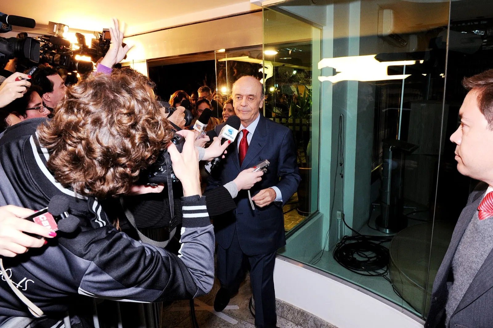 José Serra, de terno escuro e gravata vermelha, cercado por repórteres com microfones e câmeras na entrada de um estúdio.