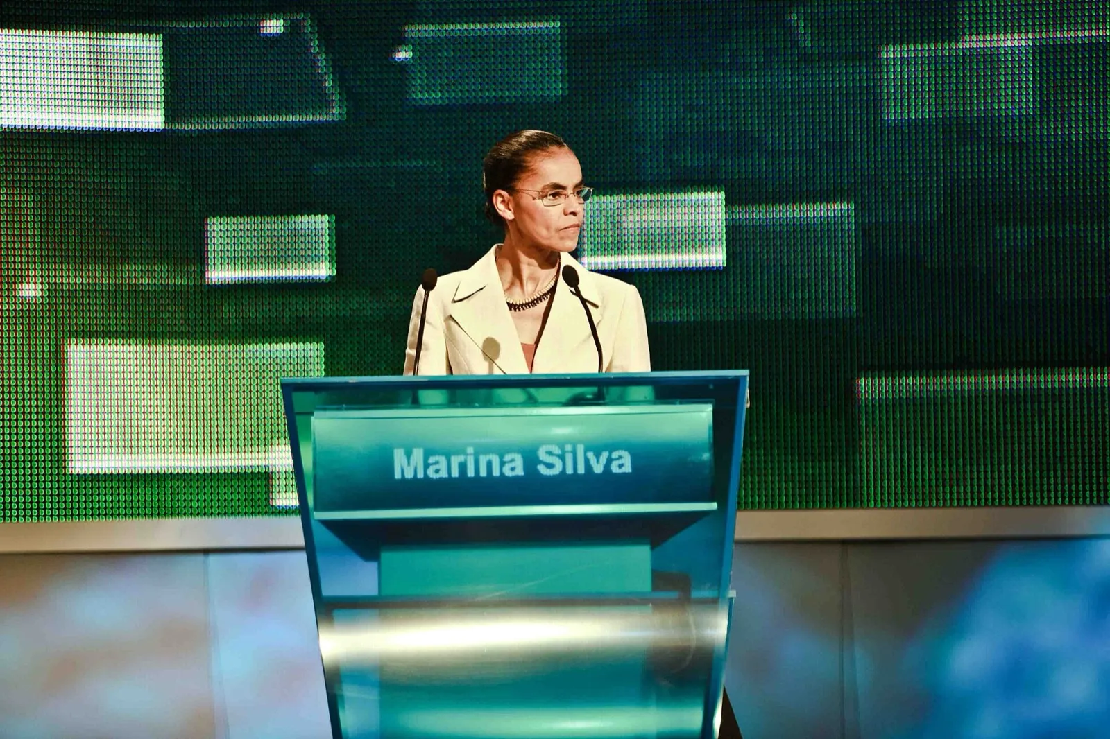 Marina Silva, de blazer claro, atrás de um púlpito com seu nome, diante de um painel verde em estúdio de televisão.