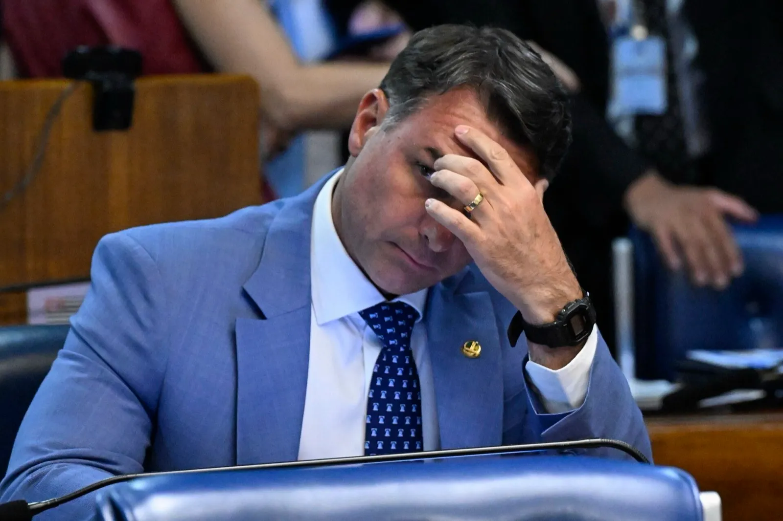 Flávio Bolsonaro, de terno azul e gravata, sentado à bancada do Plenário do Senado, com a mão na testa.